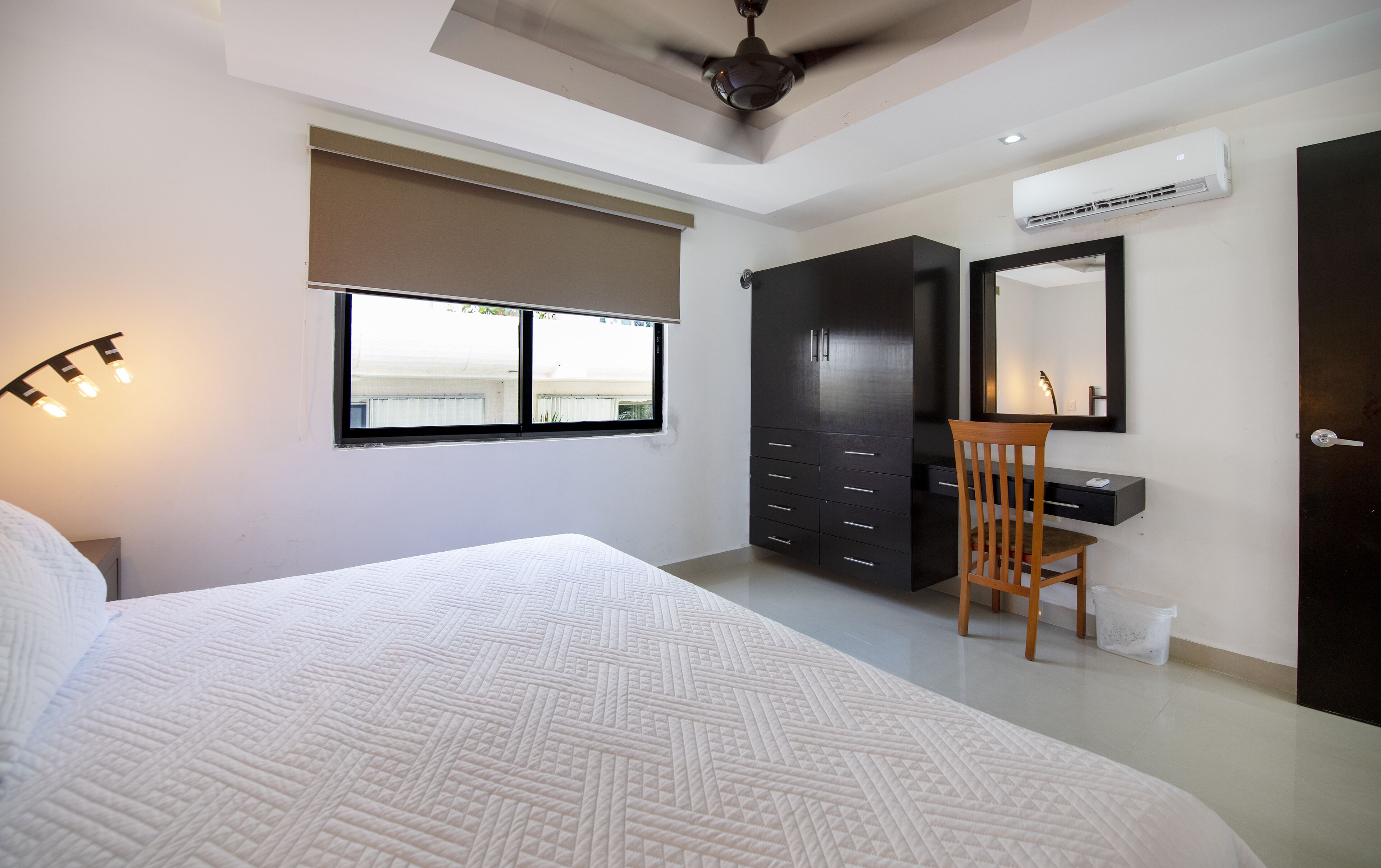 Ein Schlafzimmer mit Bett, Schreibtisch mit Stuhl, Kleiderschrank, Deckenventilator und Klimaanlage.