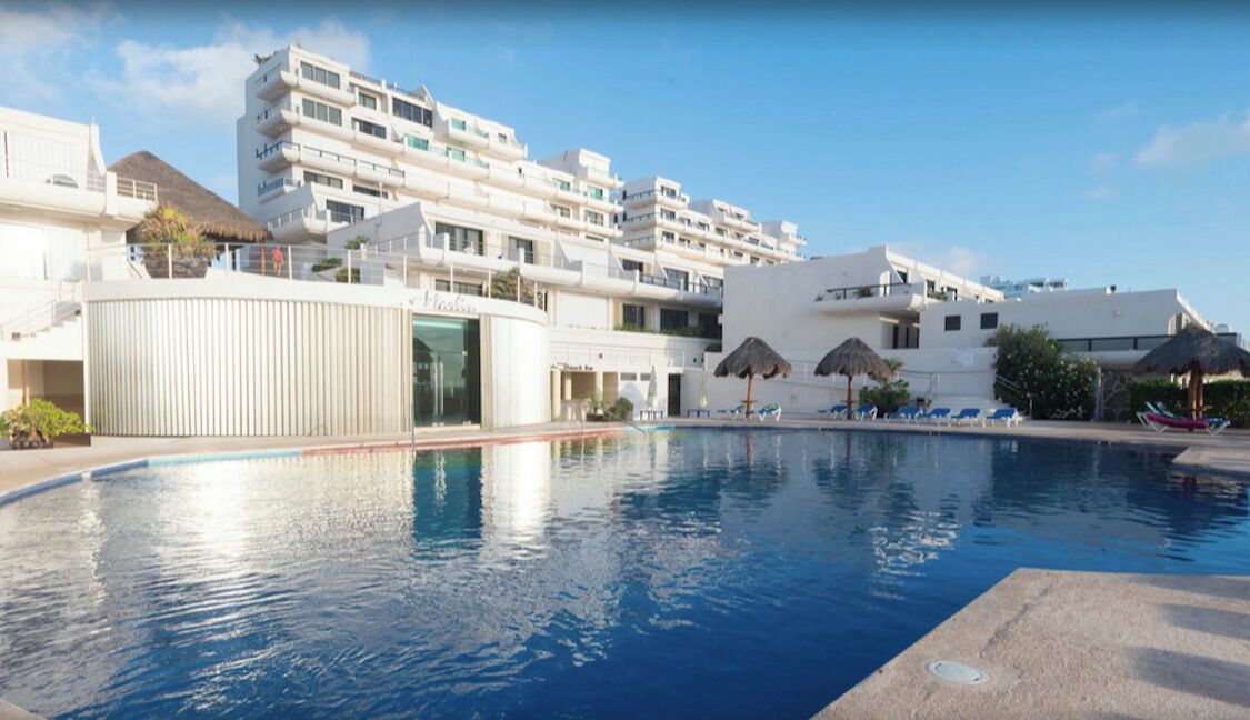 A swimming pool with a clear blue water surface, surrounded by white buildings with balconies and a clear sky.