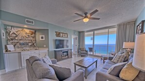 Uma sala de estar ampla com um sofá cinza, uma mesa de centro de madeira e uma TV de tela plana. O espaço possui uma grande janela com vista para o oceano e um ventilador de teto.