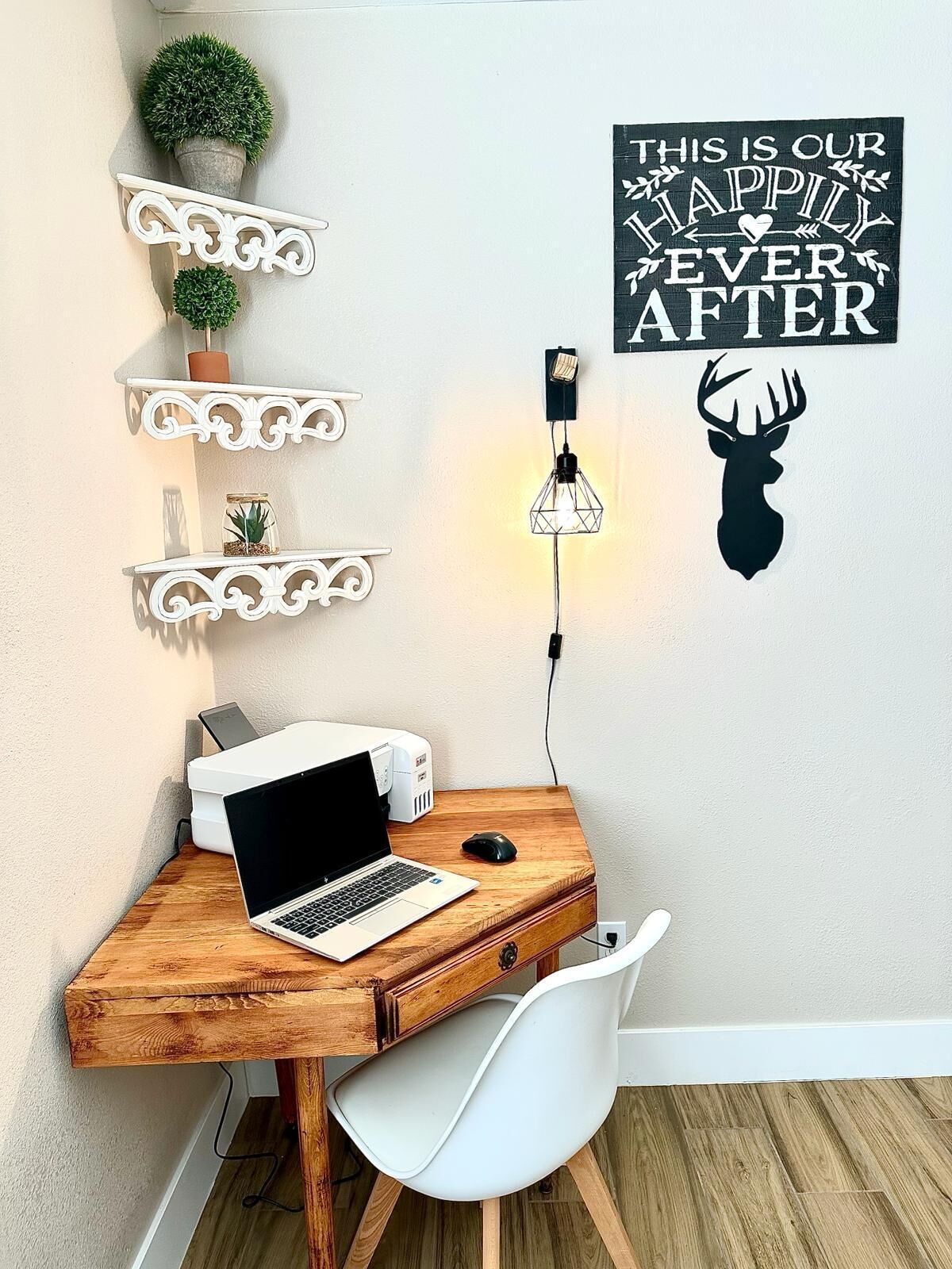 Ein Arbeitsplatz mit einem Laptop, einer Maus und einem Drucker auf einem Holztisch. Dekorative Regale mit Pflanzen und eine Wanddekoration mit der Aufschrift 'THIS IS OUR HAPPILY EVER AFTER'.