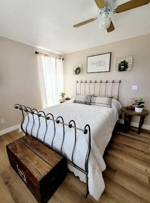 Une chambre à coucher avec un lit, un banc en bois, un ventilateur de plafond et une petite table sur laquelle est posée une plante.