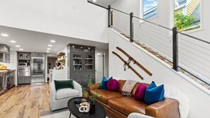 Ein geräumiger Wohnbereich mit einem braunen Ledersofa, einem weißen Sessel und einem schwarzen Couchtisch. Eine Treppe mit Metallgeländer führt auf eine obere Ebene.