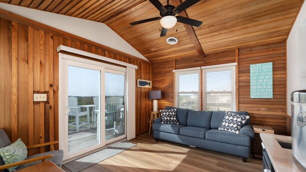 A cozy living room with wooden walls, a ceiling fan, a sofa, and a TV. - Carolina Sol: Semi-Oceanfront, Private Pool, Close to beach and Outer Banks Fishing Pier (Nags Head)