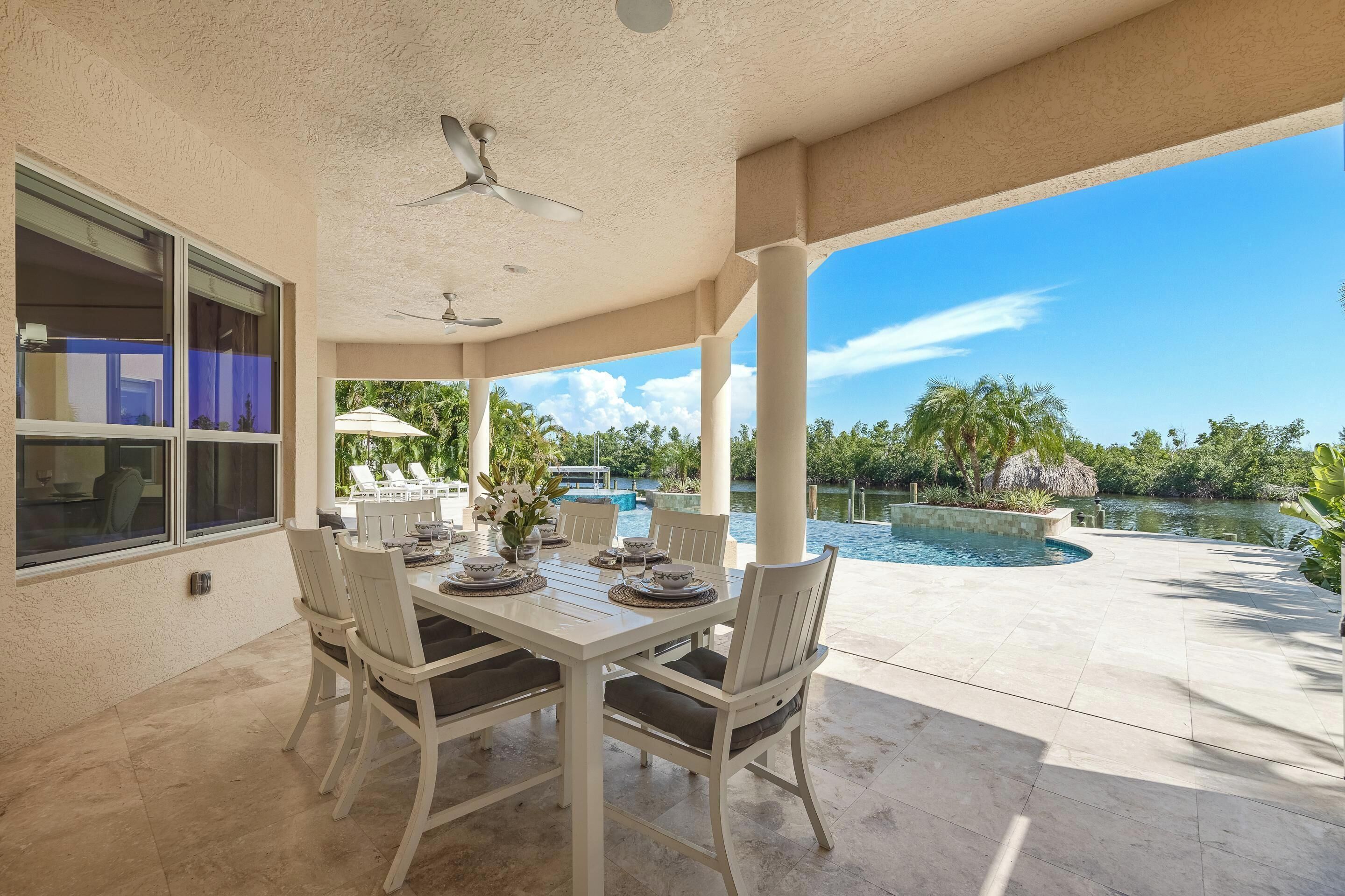 Une terrasse couverte avec une table à manger pour quatre personnes, un ventilateur de plafond et une vue sur une piscine et un paysage tropical.