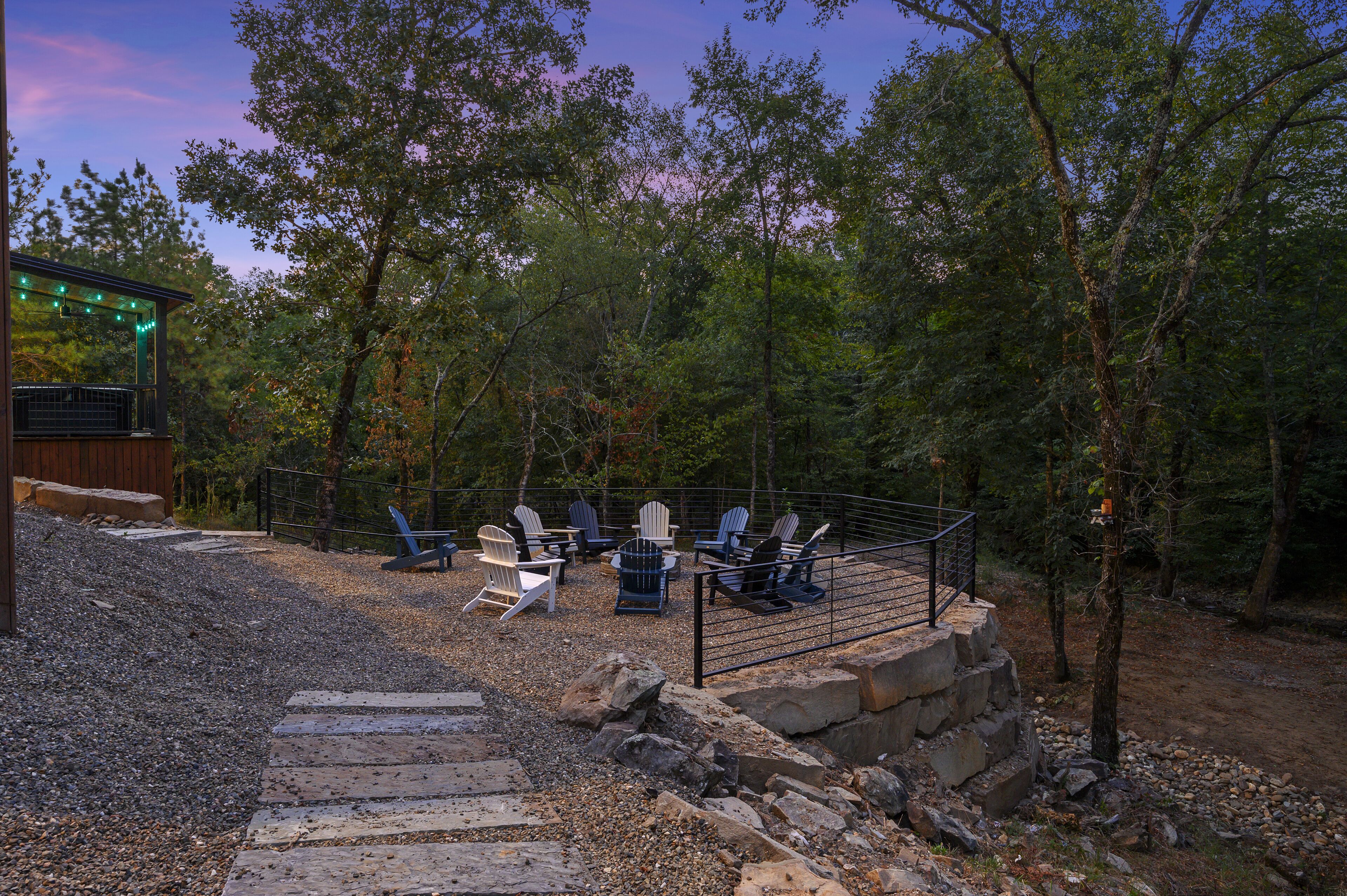 Area salotto all'aperto con poltrone in stile Adirondack e un braciere, circondata da alberi e un muro di pietra.