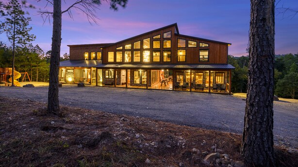 Une maison moderne en bois, à plusieurs étages, dotée de grandes fenêtres et d’une allée en gravier, et entourée d’arbres.