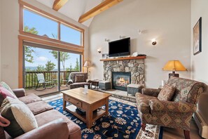 A spacious living room with a stone fireplace, a television mounted above it, and a balcony with outdoor seating.