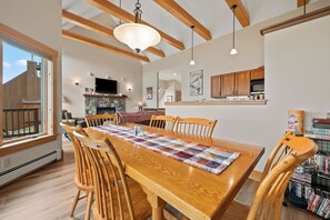 A dining area with a wooden table, chairs, and a quilt. There is a fireplace, a television, and a kitchen area with wooden cabinets. - Saddleback - Black Gnat Condo #12 (Sandy River Plantation)