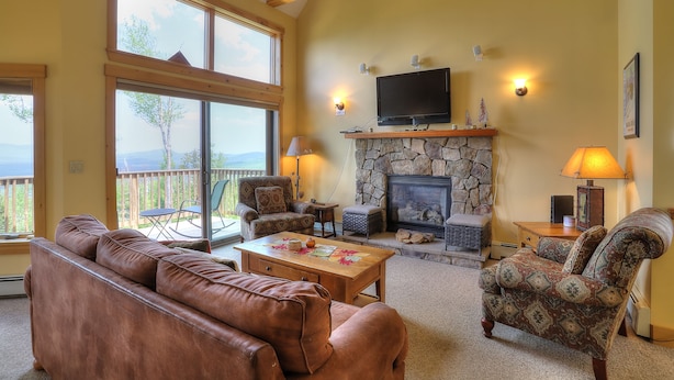 A cozy living room with a stone fireplace, comfortable seating, and large windows overlooking a deck. - Saddleback - Black Gnat Condo #12 (Sandy River Plantation)