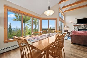 A spacious dining area with a wooden table and chairs, large windows offering a view of trees and mountains, a stone fireplace, and a television mounted on the wall. - Saddleback - Black Gnat Condo #12 (Sandy River Plantation)
