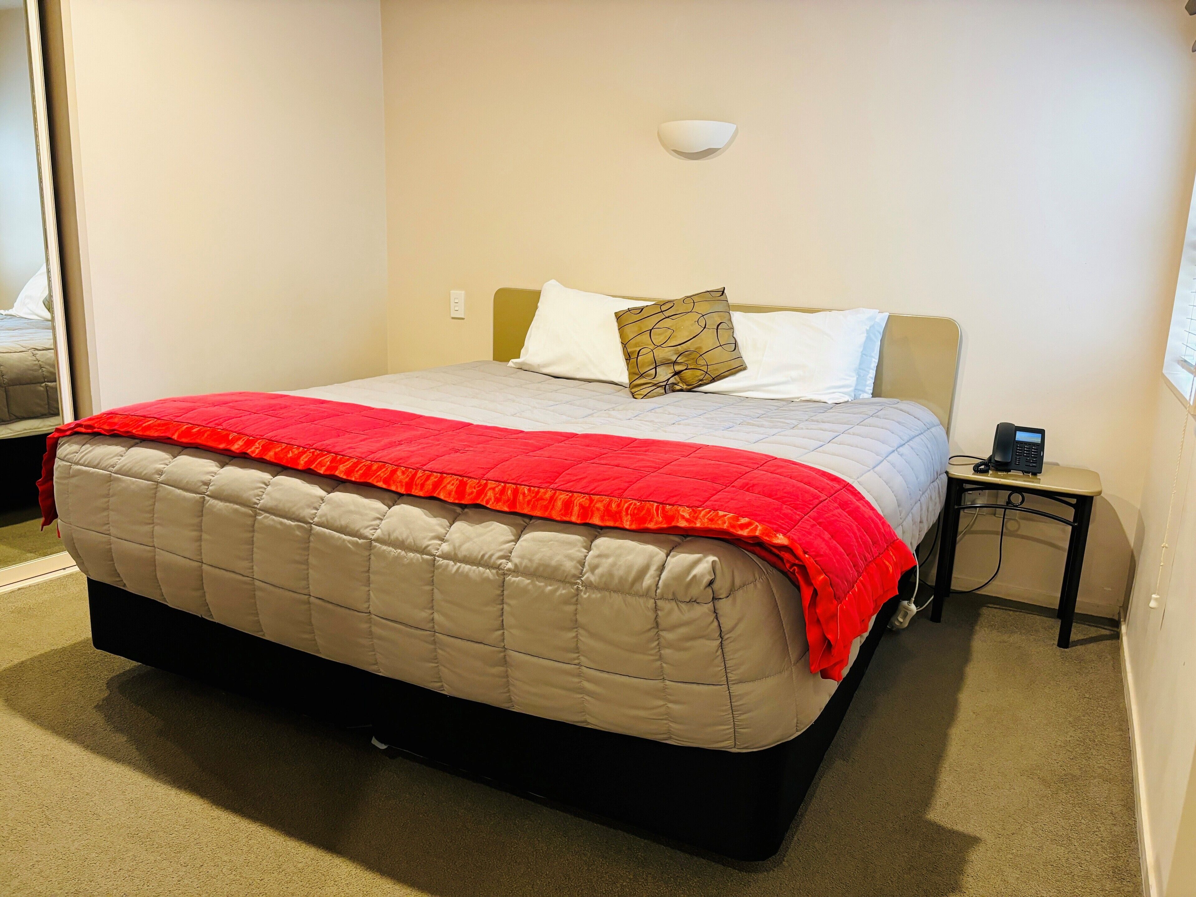 A neatly made bed with a red and beige quilt, a bedside phone, and a wall-mounted light fixture.
