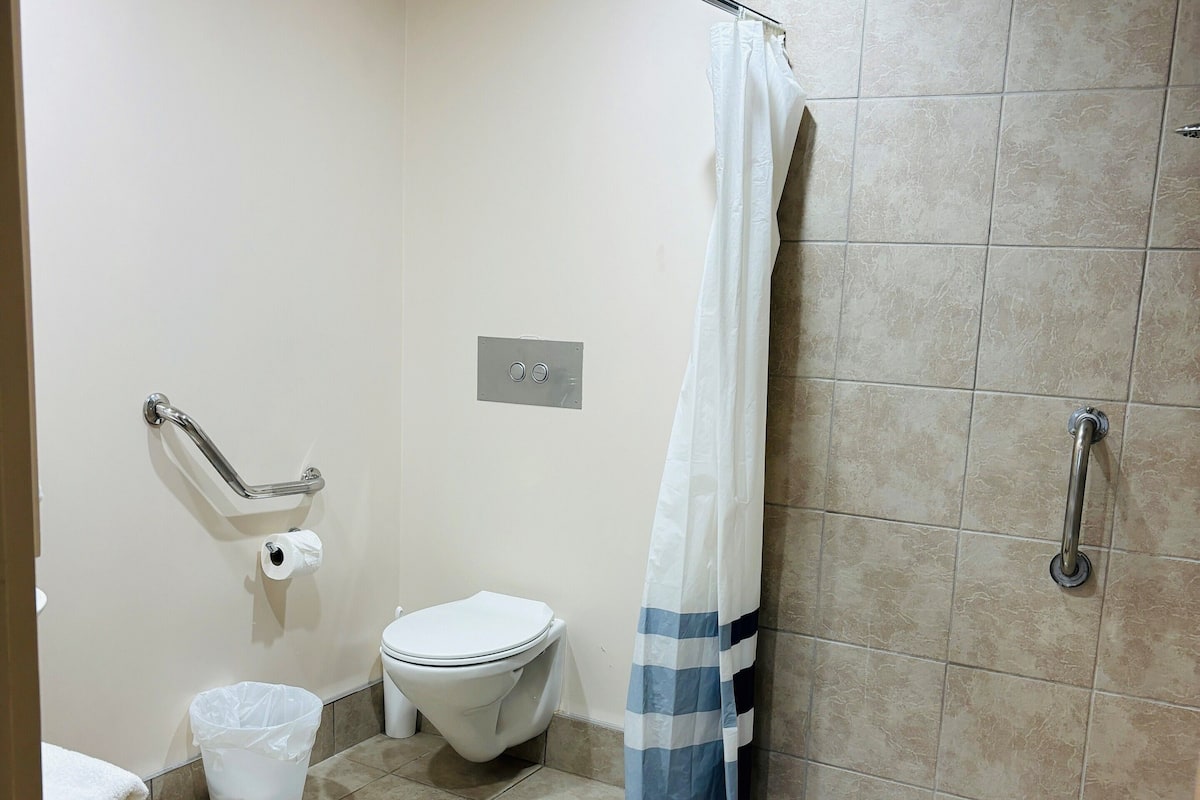 A bathroom with a toilet, a shower with a curtain, a grab bar, and a trash bin.