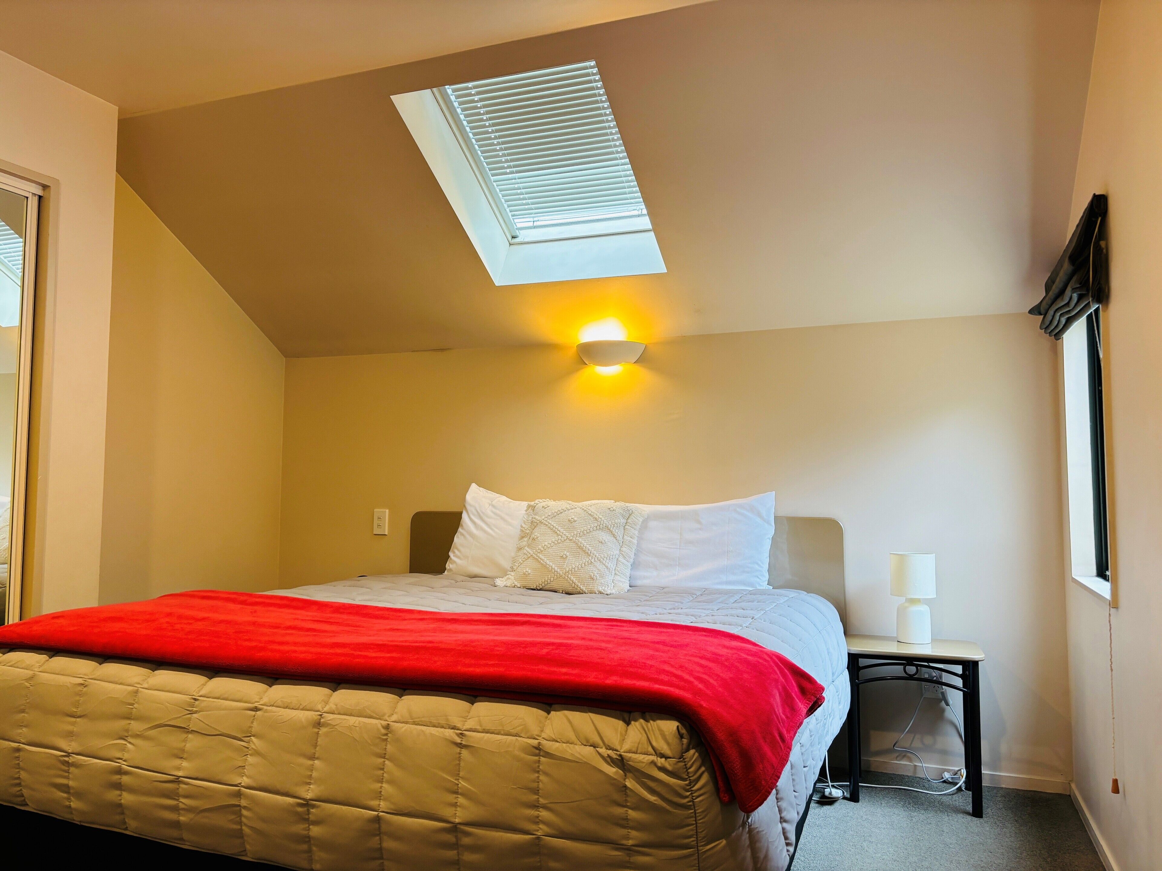 A bedroom with a bed, a nightstand, a lamp, a window with blinds, and a skylight.
