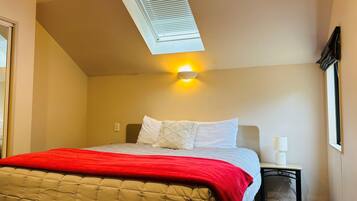 A bedroom with a bed, a nightstand, a lamp, a window with blinds, and a skylight.
