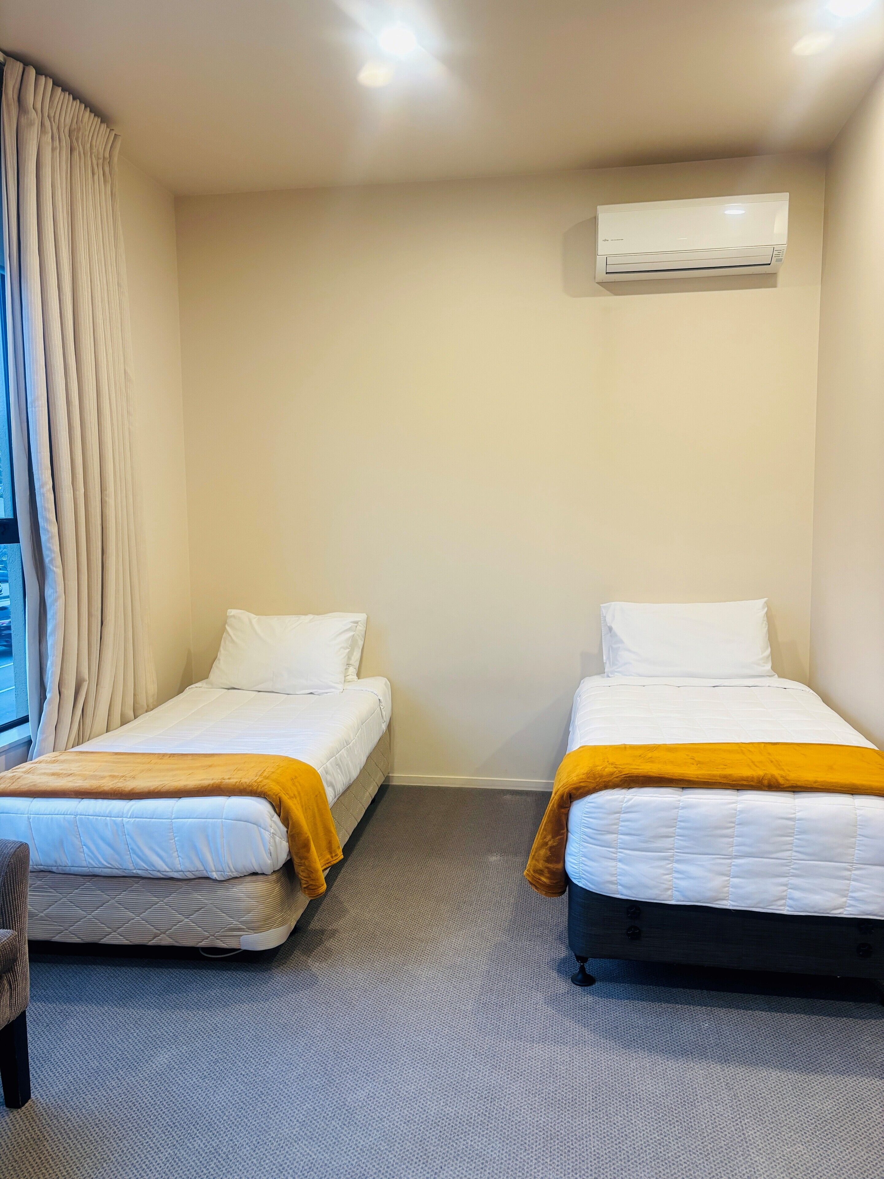Quarto de hotel com duas camas, uma janela com cortinas, ar condicionado e piso coberto de carpete.