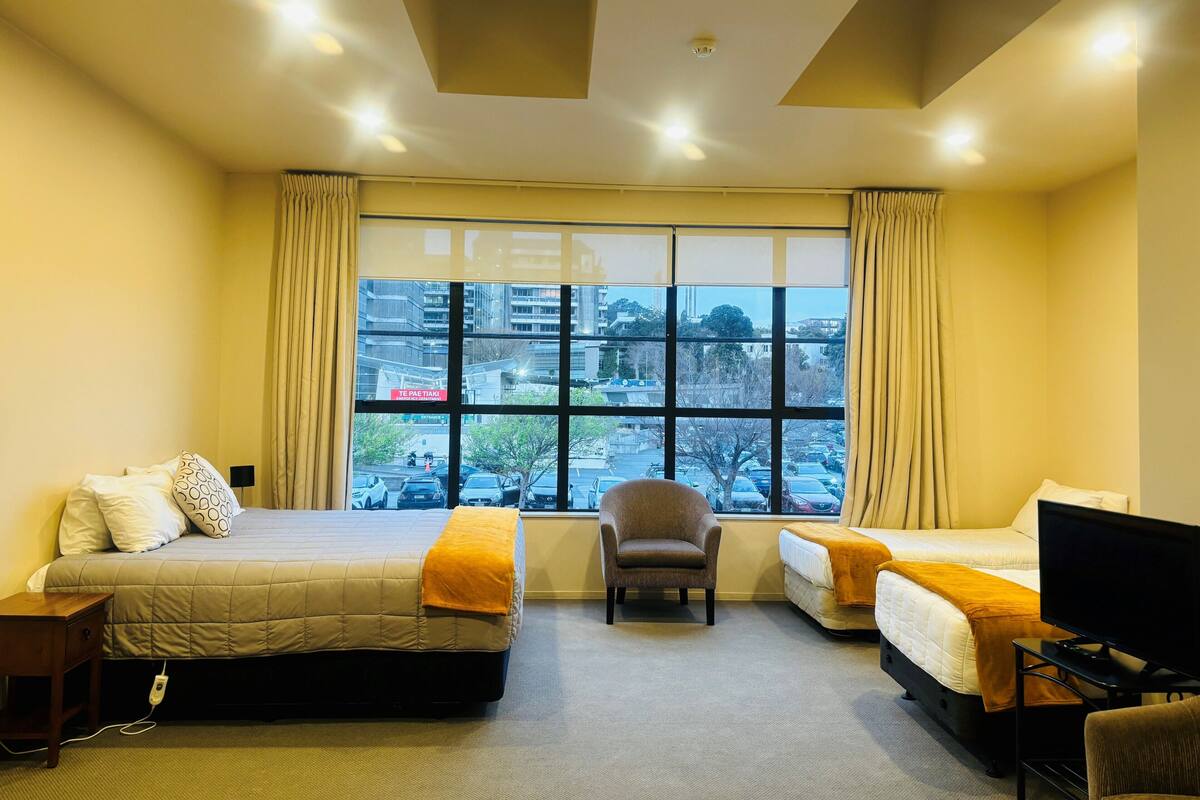 A hotel room with two beds, a chair, a TV, and a large window with a city view.
