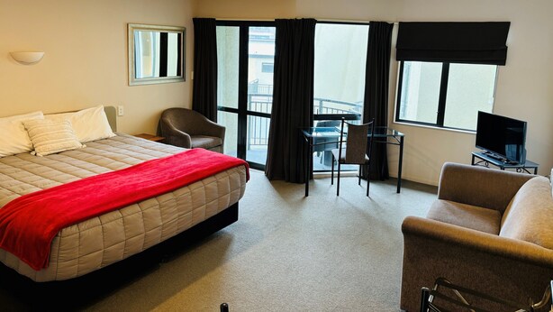 Een hotelkamer met een bed, een bureau met stoel, een televisie en grote ramen met gordijnen.