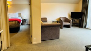 Hotelkamer met een bed, een zithoek met een tafel en stoelen, een televisie op een standaard, en een deur naar een andere kamer.