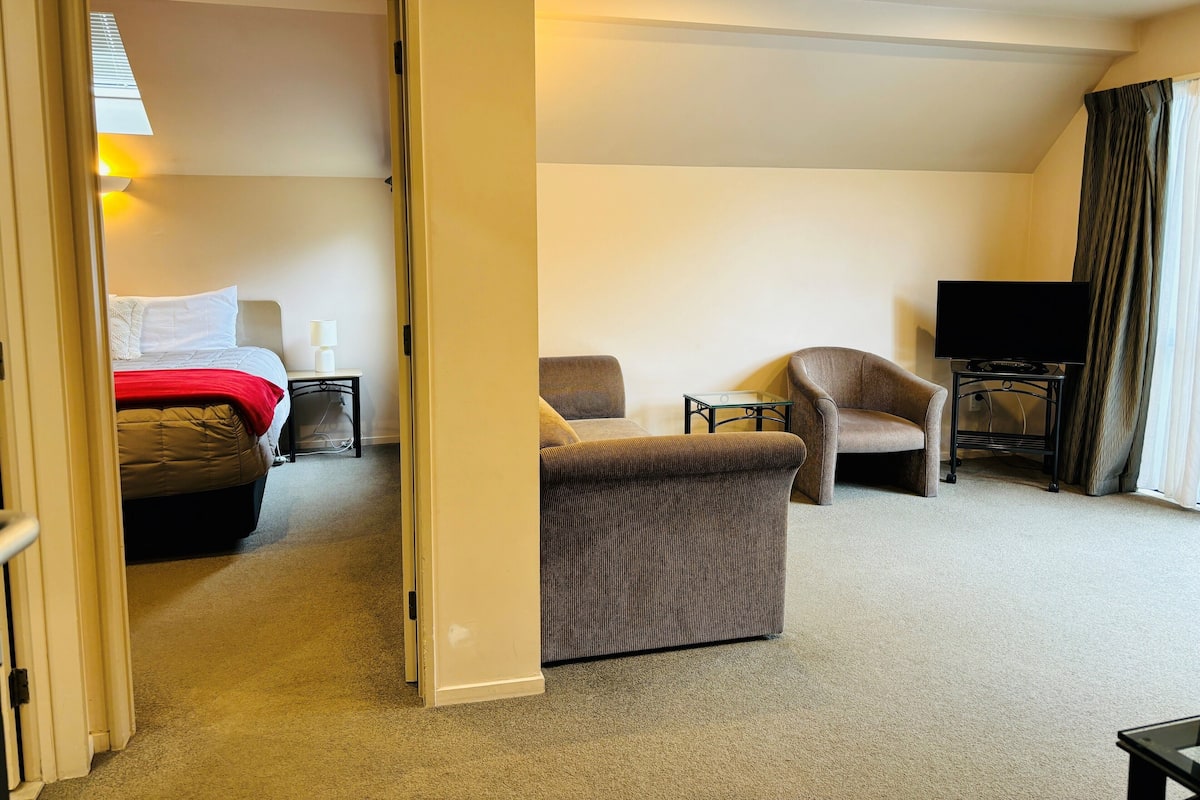 A hotel room with a bed, a sitting area with a table and chairs, a television on a stand, and a door leading to another room.