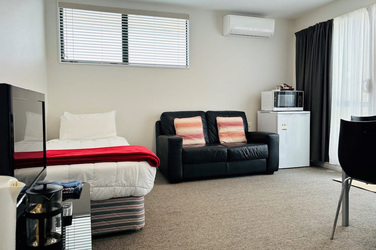 A hotel room with a bed, a black sofa with two cushions, a small kitchenette, and a TV.