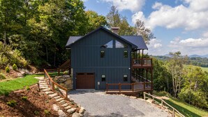 A two-story house with a balcony, surrounded by trees and a gravel driveway. - Blue Heaven - Long Range Views, Hot Tub, Luxury Home, Near Blowing Rock! (Boone)