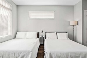 A room with two beds, a window with blinds, a bedside lamp, and a door.