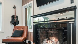 A living room with a fireplace, a leather chair, a guitar, and a television. - Luxe | Hot Tub | Firepit | Heat Floors | EV | Walk2Matthews (Matthews)