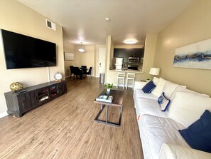 Una sala de estar con televisor de pantalla plana, mueble de madera oscura, sofá blanco con cojines azules y una mesa de centro.