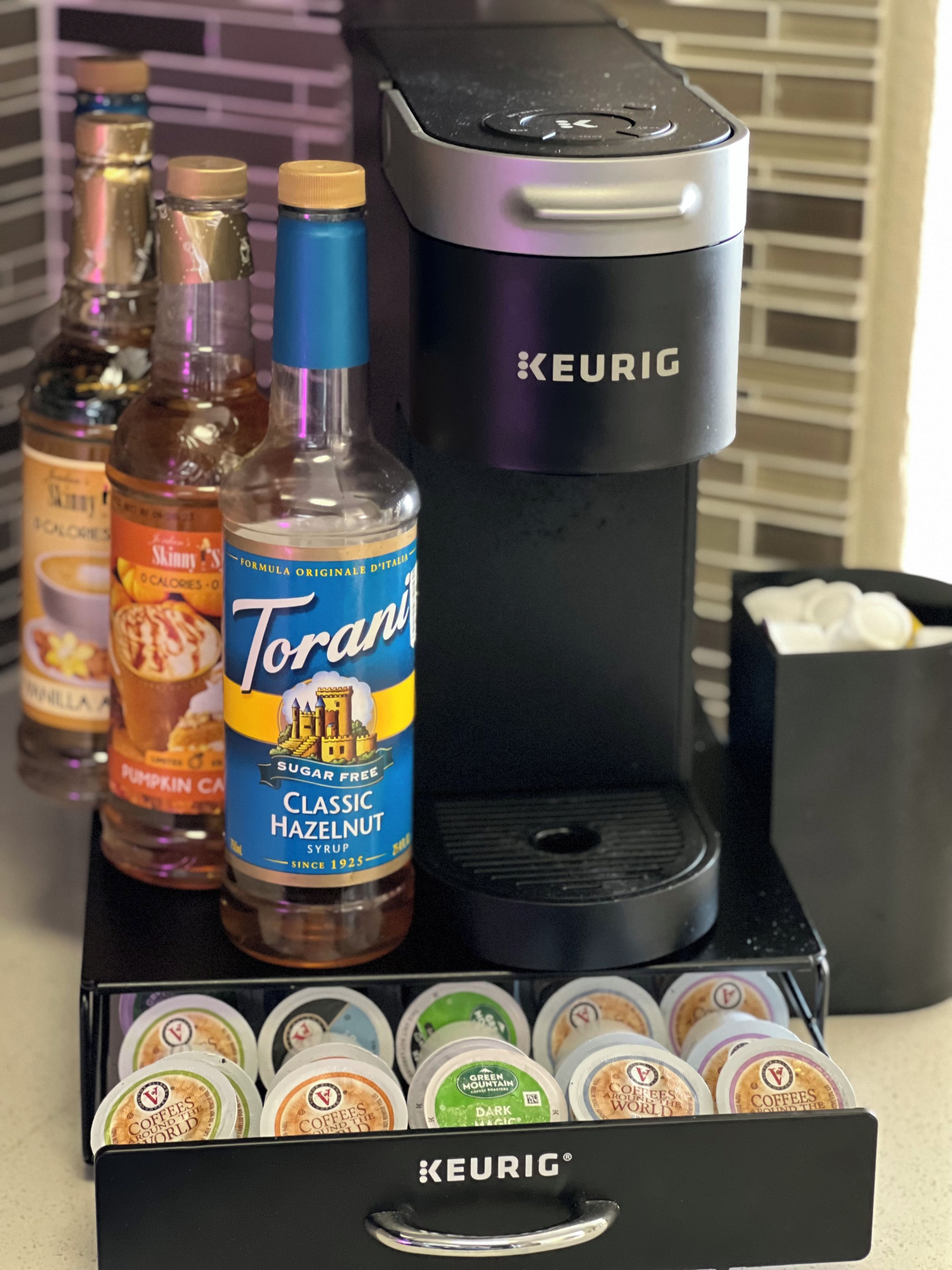 A coffee maker, Torani syrup bottle, and coffee pods on a countertop.