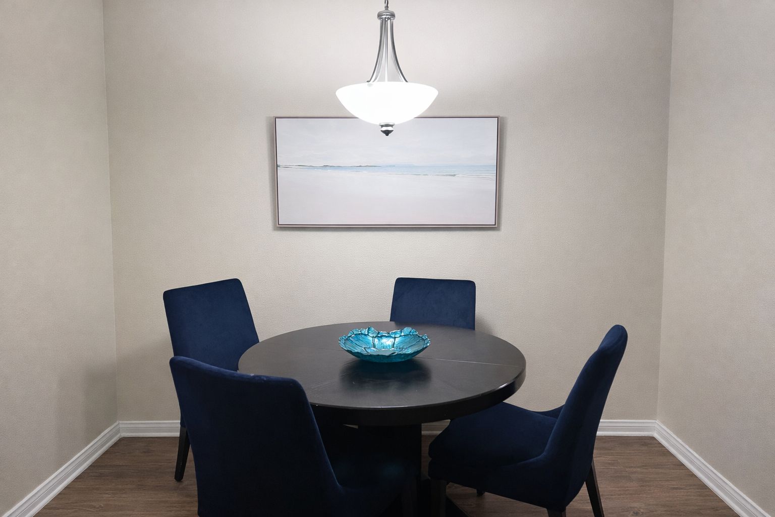 A dining area with a round table, four chairs, a glass bowl centerpiece, a hanging light fixture, and a framed picture on the wall.