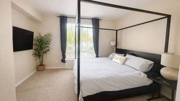 A four-poster bed with a dark frame, a flat-screen TV mounted on the wall, a potted plant, and a bedside table with a lamp.
