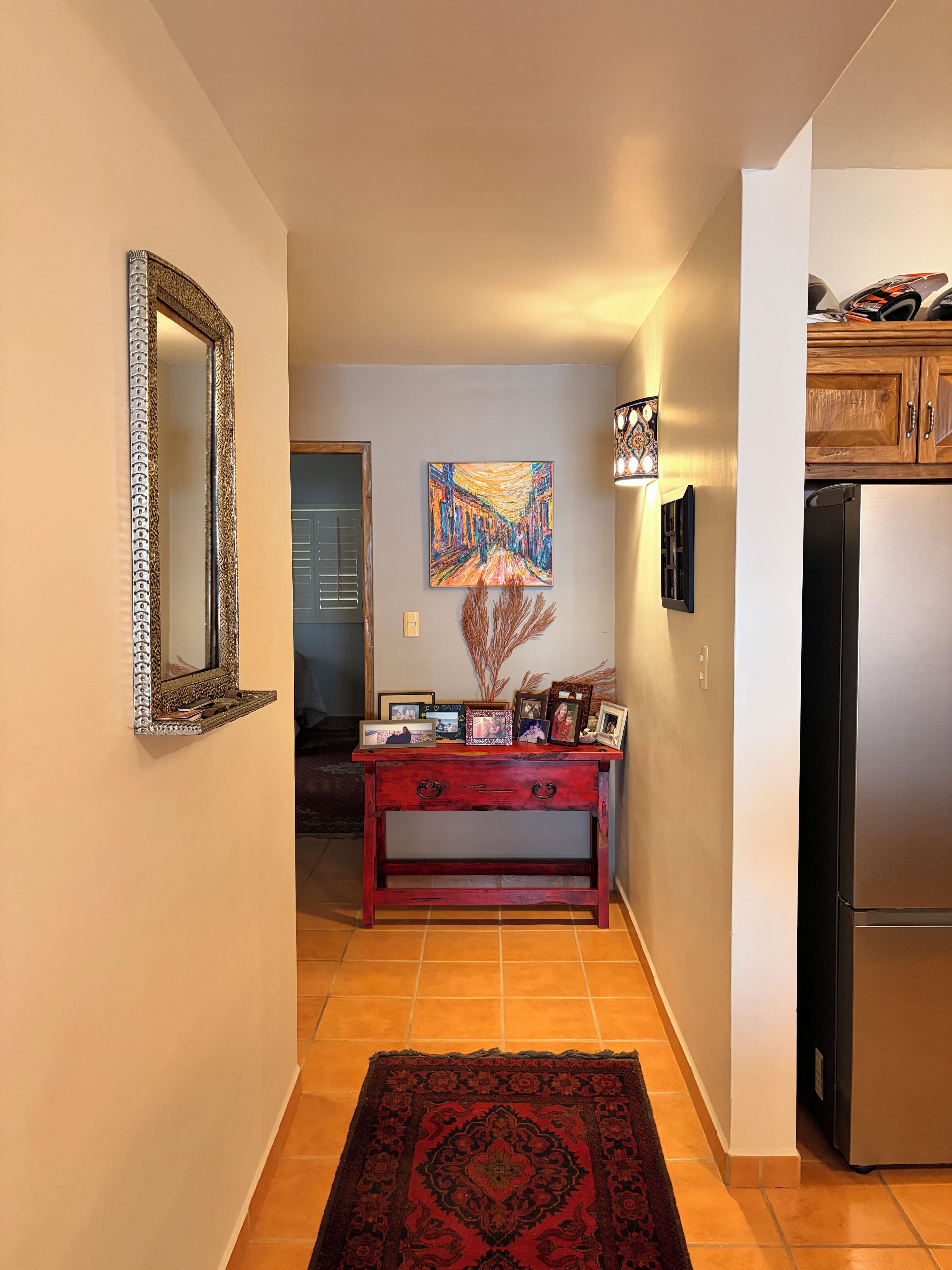 A hallway with a wooden console table, a mirror, a painting, and a refrigerator.