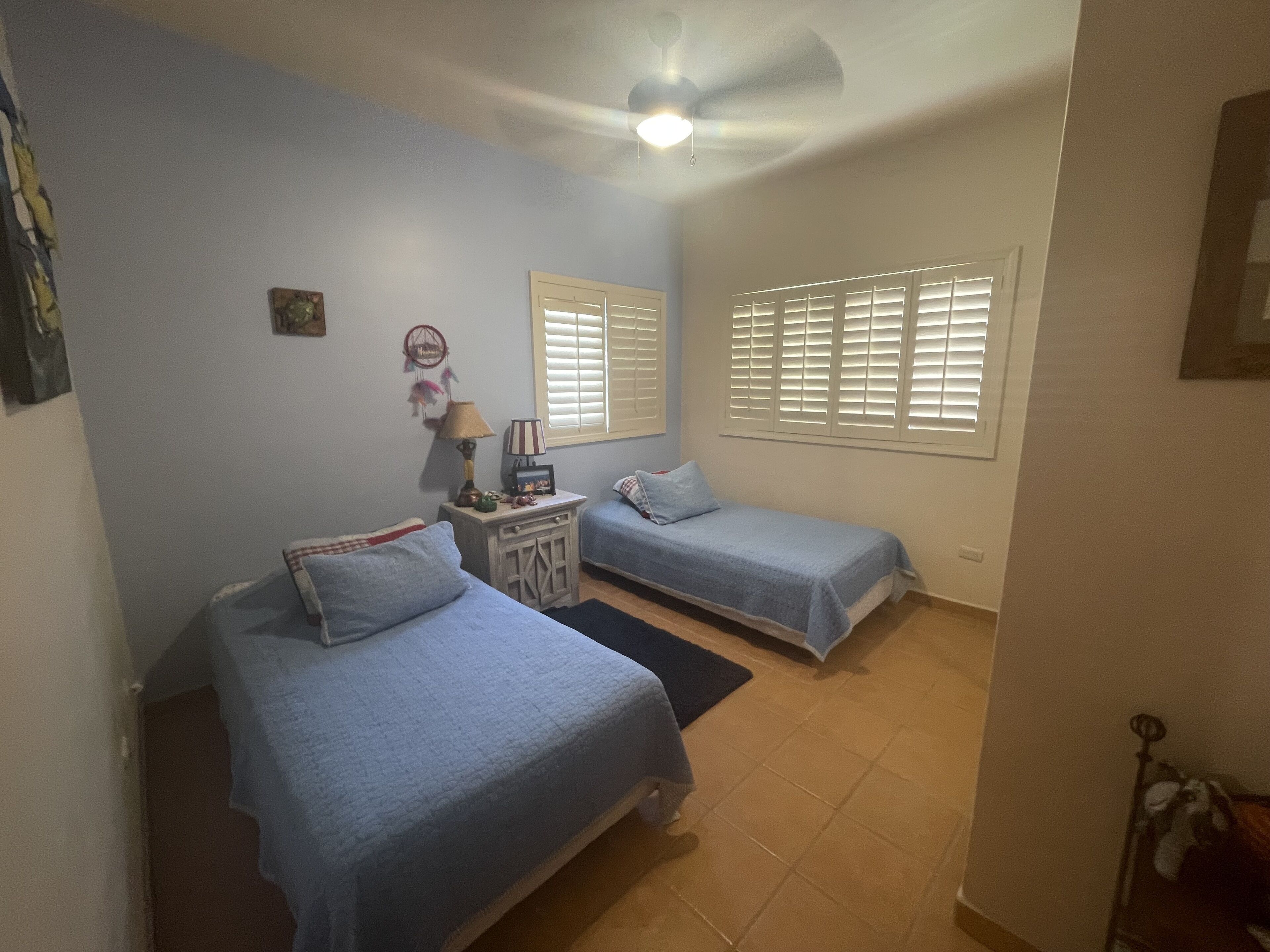 Une chambre avec deux lits simples, une table de chevet, un ventilateur de plafond et une fenêtre avec des stores.