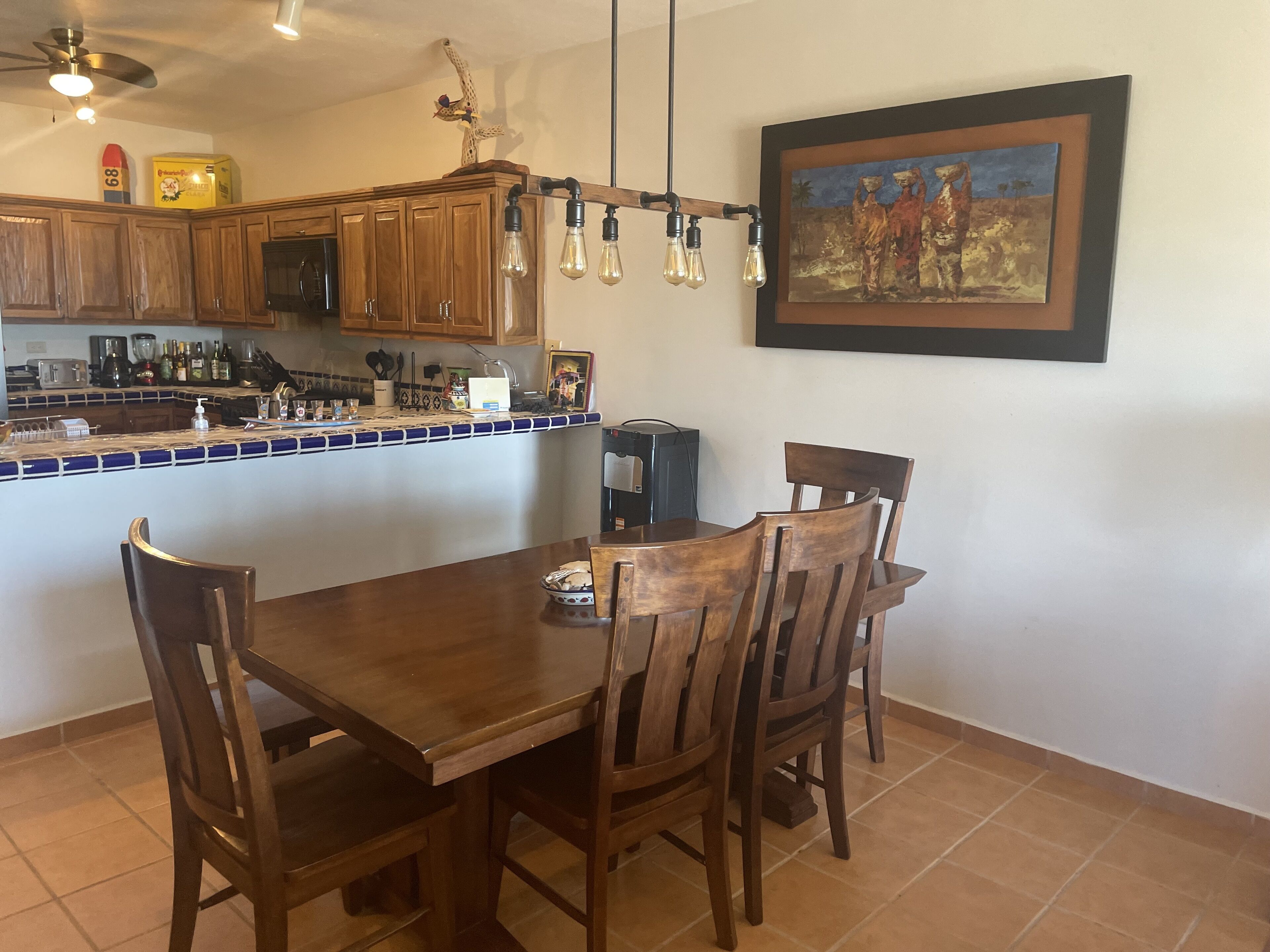 Un coin repas avec une table et des chaises en bois, une cuisine avec des meubles en bois et un dosseret carrelé, et un ventilateur de plafond.