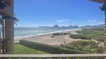 A beach view from a balcony with a railing, clear sky, and distant mountains.