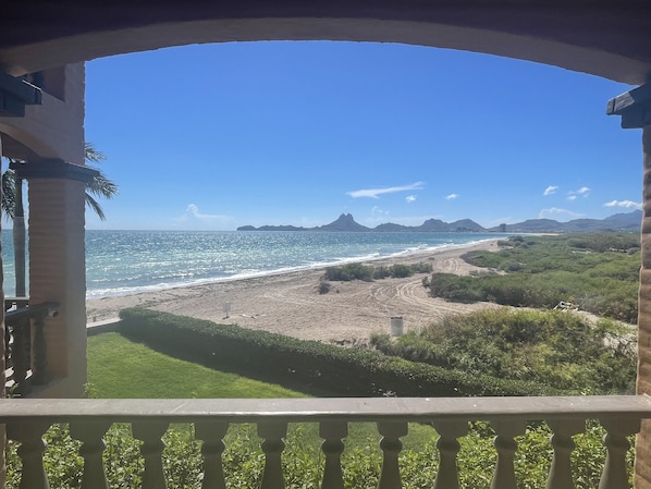 A beach view from a balcony with a railing, clear sky, and distant mountains.