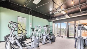 Un gimnasio con varias máquinas de ejercicio, una pared verde y grandes ventanales con vistas a un balcón.