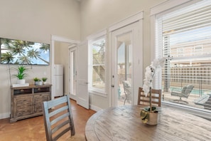 A dining area with a round wooden table, chairs, a white cabinet with plants, and a large window with blinds.