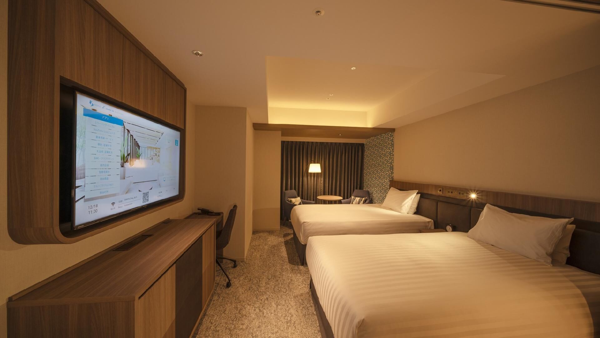 A hotel room with two beds, a desk, a TV mounted on the wall, and a curtain backdrop.