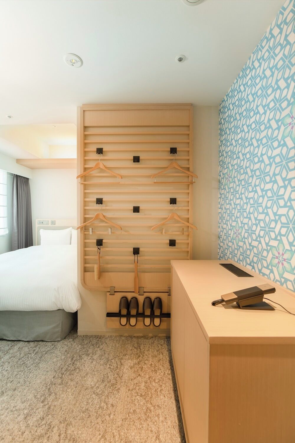 A hotel room with a bed, a wooden wardrobe with hanging space, a desk with a chair, and a patterned wall.