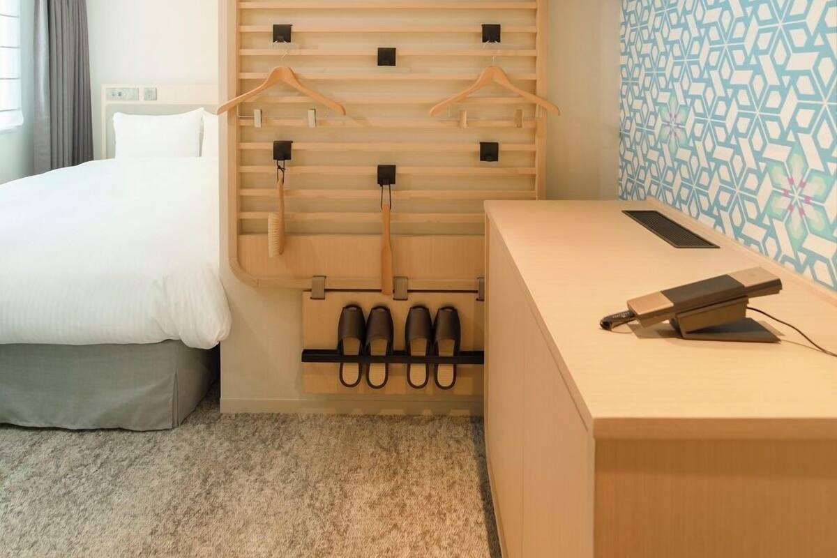 Une chambre d’hôtel comprenant un lit, une armoire en bois avec espace pour suspendre les vêtements, un bureau avec une chaise et un mur décoré d’un motif.