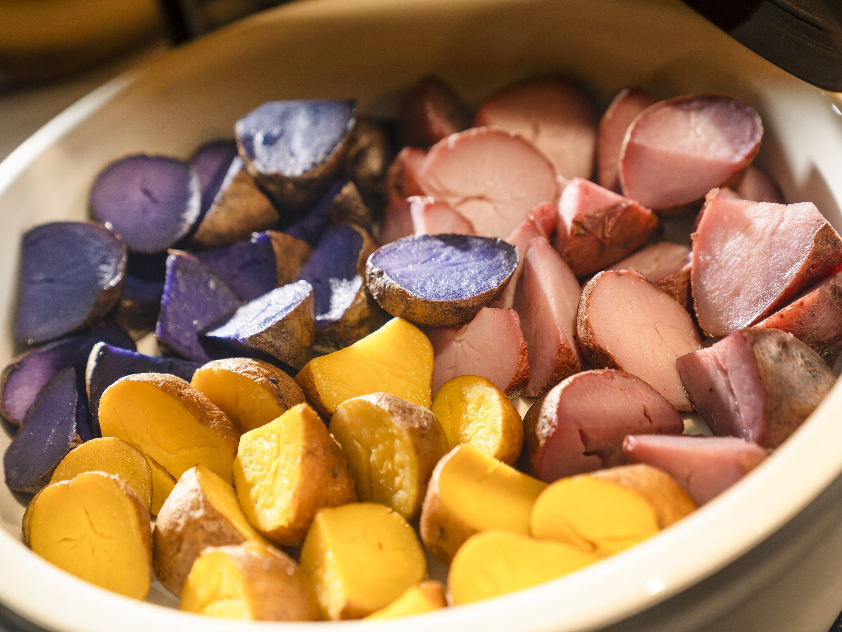 A bowl containing sliced purple potatoes, yellow potatoes, and ham.
