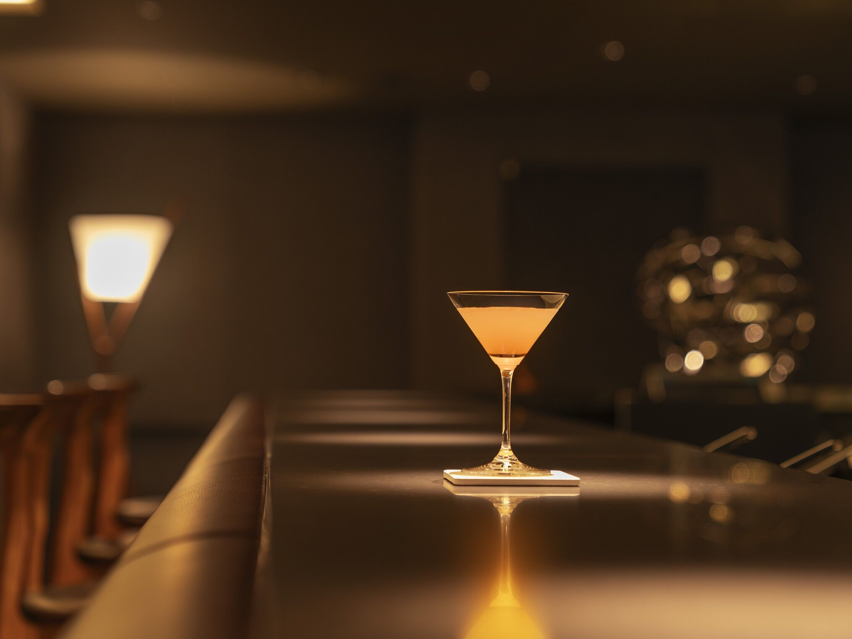 Un bar avec un seul verre à martini posé sur une surface réfléchissante, une lampe et un arrière-plan flou.