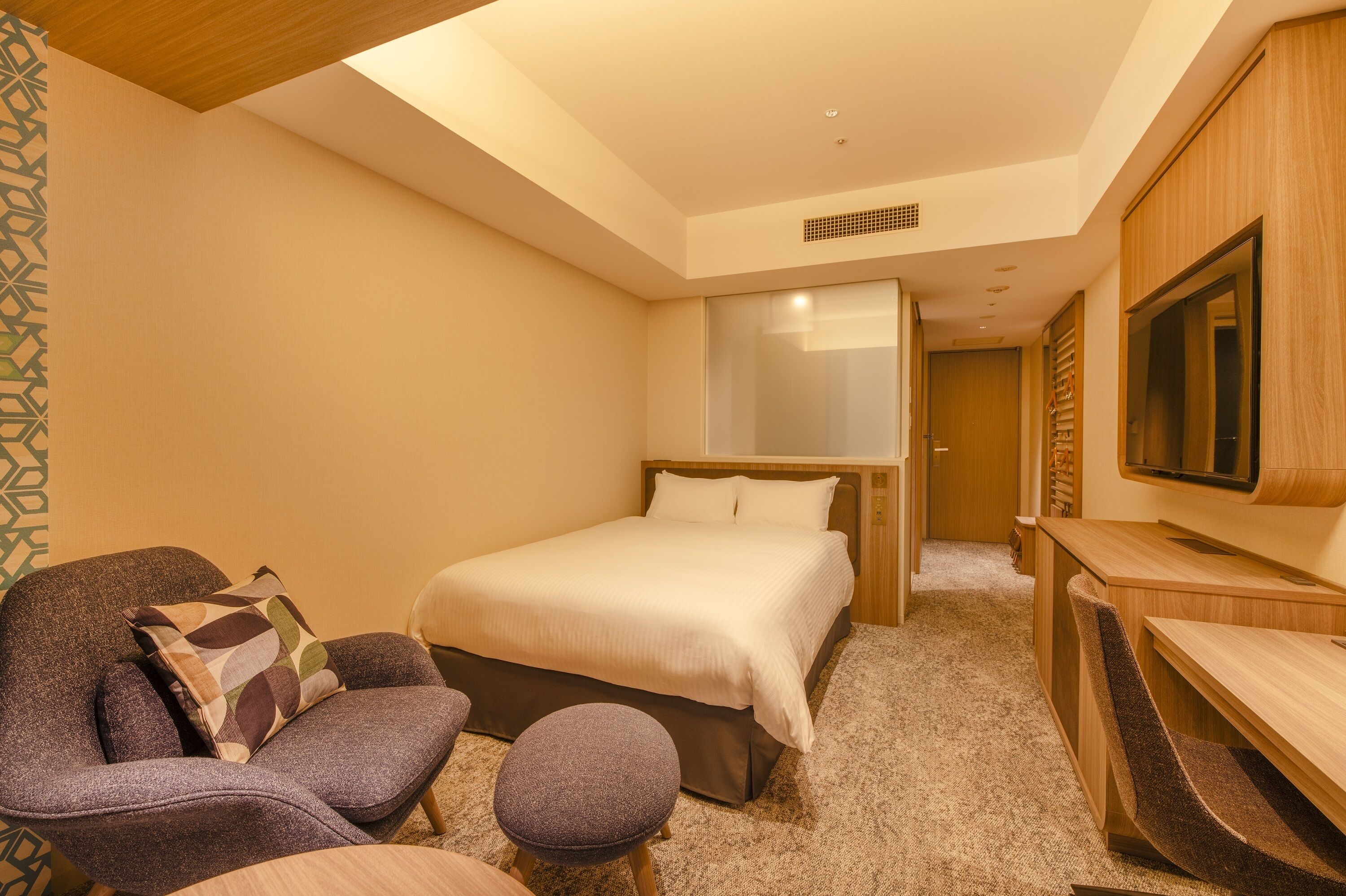 A hotel room with a bed, a seating area with a chair and ottoman, a desk, and a television.