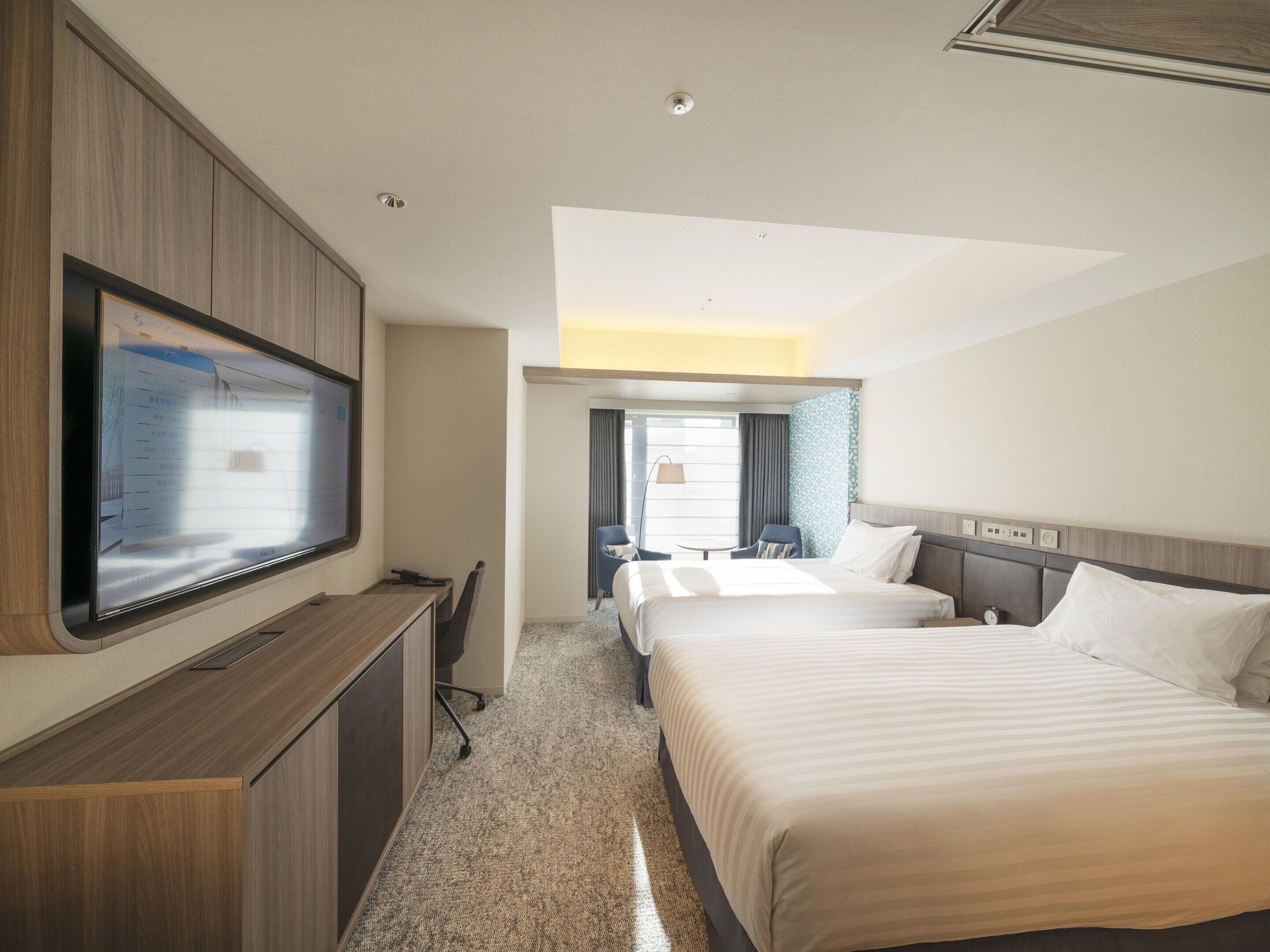A hotel room with a large flat-screen TV mounted on the wall, a desk with a chair, two beds, and a small table with a chair.