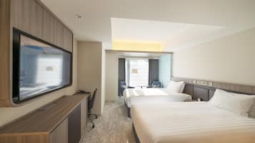 A hotel room with a large flat-screen TV mounted on the wall, a desk with a chair, two beds, and a small table with a chair.