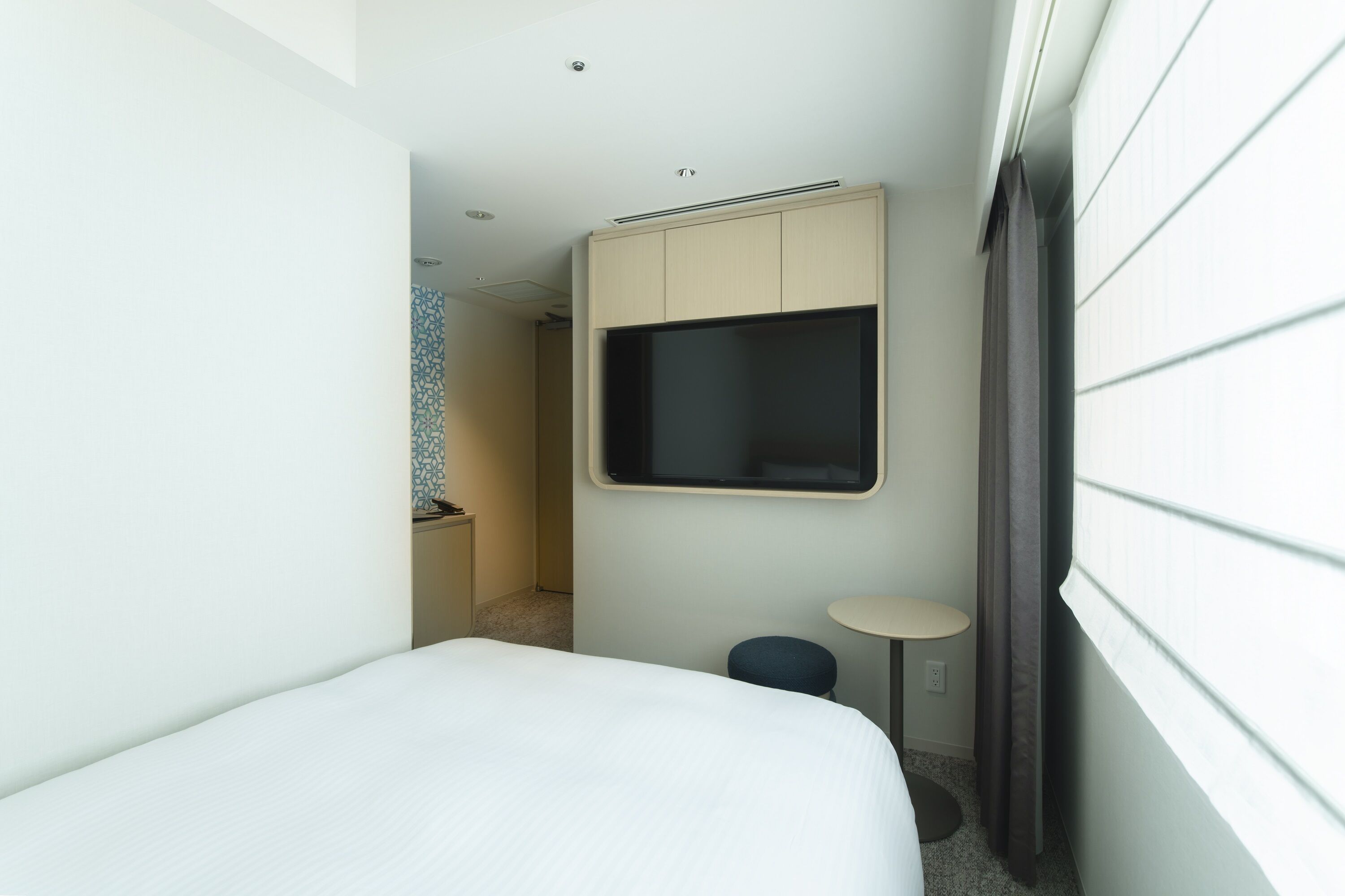 A narrow hotel room with a bed, a small round table, a TV mounted on the wall, and a window with blinds.