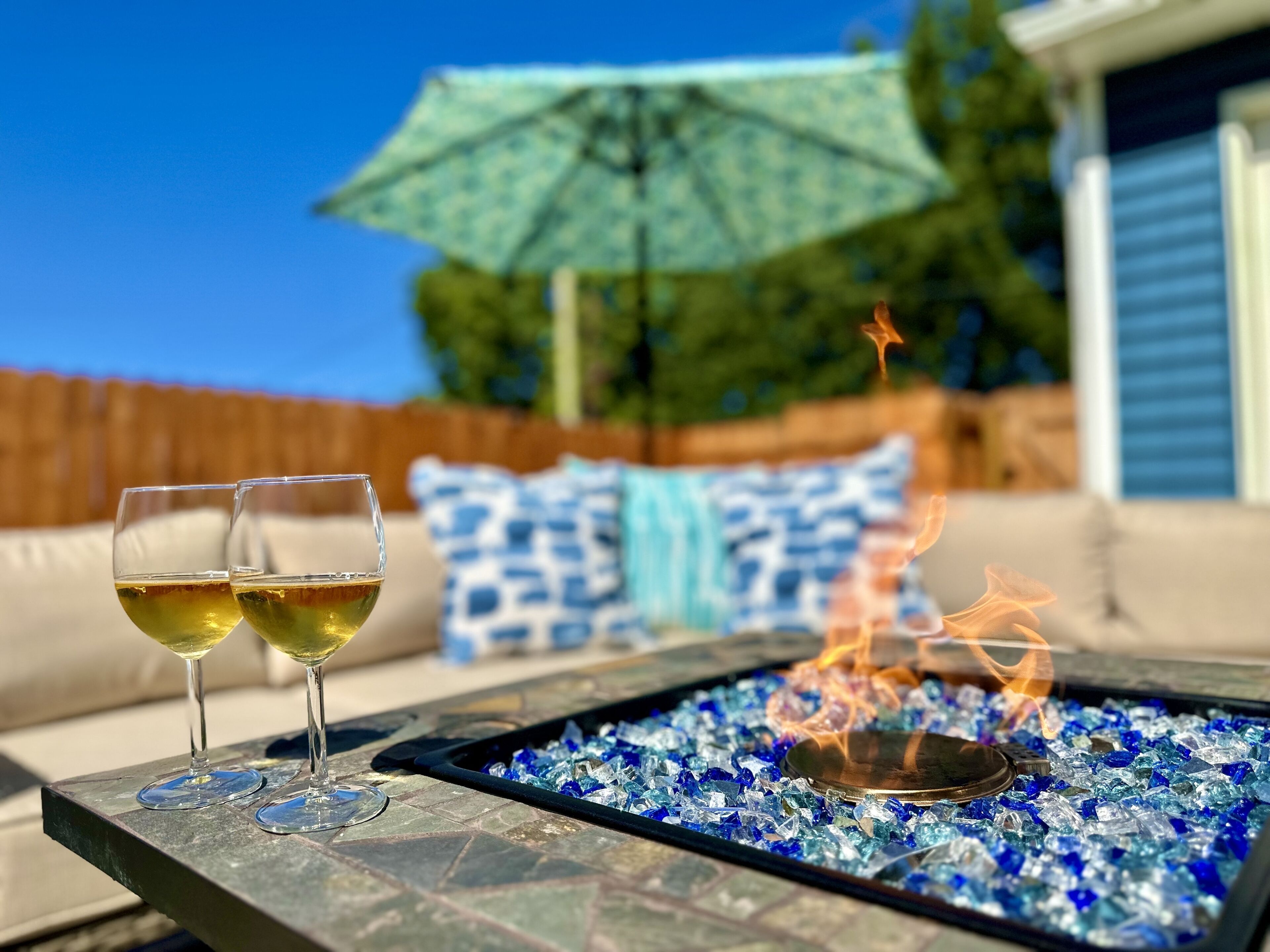 Un patio con dos copas de vino blanco, una chimenea decorativa con vidrio azul y un paraguas azul.