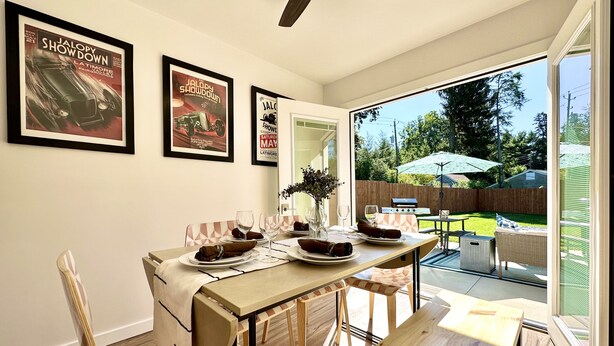 Un coin repas avec une table dressée pour quatre personnes, des affiches encadrées au mur et une vue sur le jardin à travers une porte vitrée.