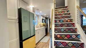 Une cuisine moderne avec des meubles blancs, un réfrigérateur noir et jaune, et un four à micro-ondes. Un escalier aux motifs colorés mène à l’étage supérieur.